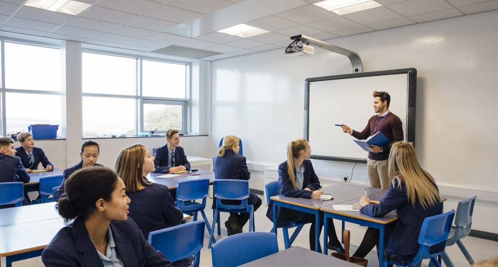 Teacher engaging with students in a modern classroom with digital whiteboard and blue desks